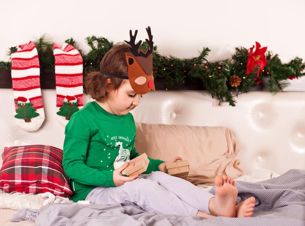 Kleines süßes Mädchen in lustiger Hirschmaske im Weihnachtspyjama, das auf dem Bett sitzt und Geschenkboxen zu Weihnachten bekommt