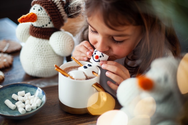 Kleines süßes Mädchen isst einen mit Zuckerguss verzierten Marmmello-Schneemann und trinkt Kakao