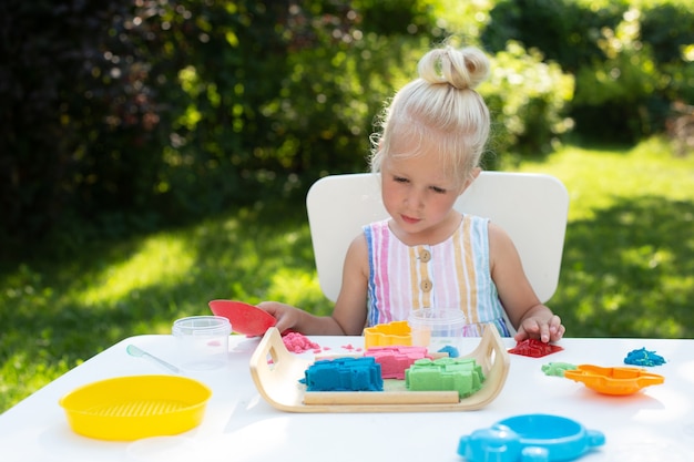 Kleines süßes Mädchen mit blonden Haaren, das am Sommertag mit buntem kinetischem Sand draußen im Hinterhof spielt. Hobby, Freizeitbeschäftigung, Basteln, Modellbau und kreatives Hobbykonzept