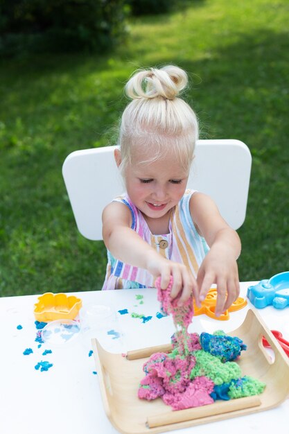 Kleines süßes Mädchen mit blonden Haaren, das am Sommertag mit buntem kinetischem Sand draußen im Hinterhof spielt. Hobby, Freizeitbeschäftigung, Modellbau, Basteln. Vertikales Format.