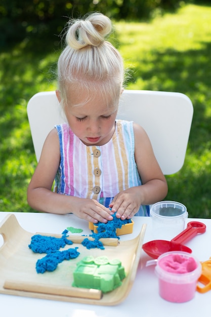 Kleines süßes Mädchen mit blonden Haaren, das am Sommertag mit kinetischem Sand draußen im Hinterhof spielt