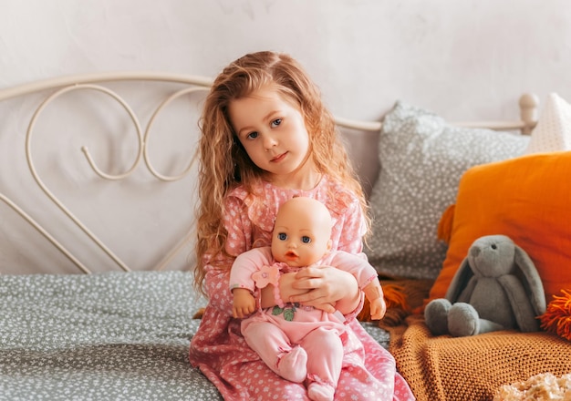 Kleines süßes Mädchen mit einer Puppe sitzt auf einem Bett in einem Kinderzimmer. Fröhliche Kindheit, fröhlicher Lebensstil, Spiele, gemütlicher Zeitvertreib, Hobbykonzept