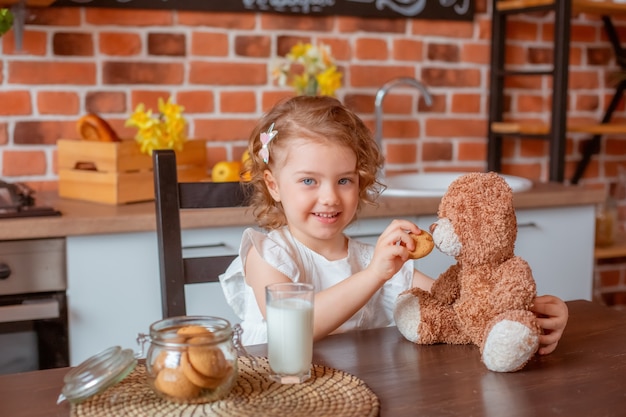 Kleines süßes Mädchen mit Teddybär-Trinkmilch zum Frühstück in der Küche zu Hause