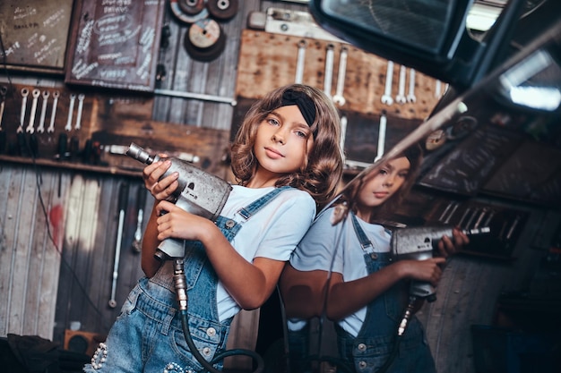 Kleines süßes Mädchen posiert mit pneumatischer Bohrmaschine für Fotografen in der Autowerkstatt.