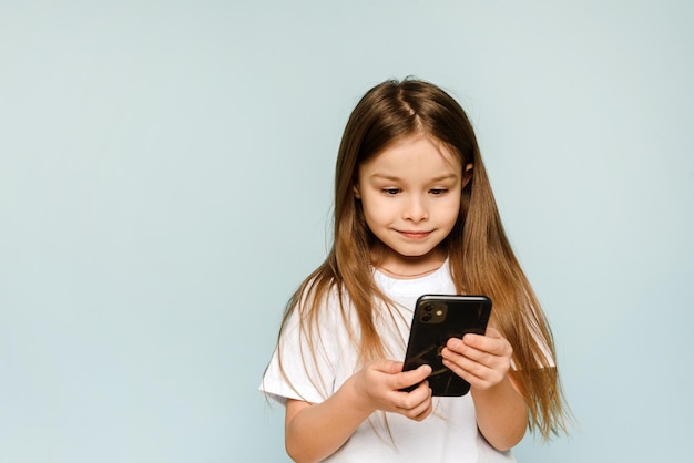 Kleines süßes Mädchen schaut auf den Bildschirm des Telefons, lächelt und genießt das Gadget auf blauem Hintergrund