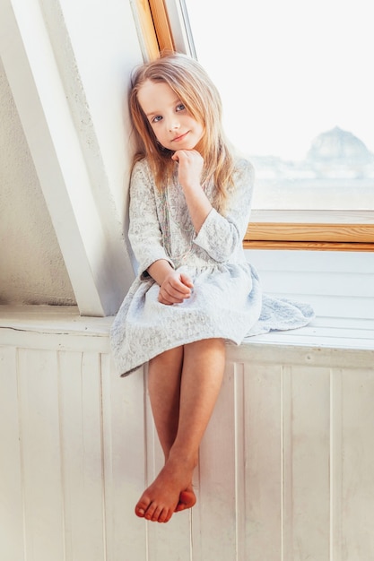 Kleines süßes süßes lächelndes Mädchen im weißen Kleid, das auf der Fensterbank im hellen Wohnzimmer zu Hause drinnen sitzt. Kindheit Schulkinder Jugend entspannen Konzept.