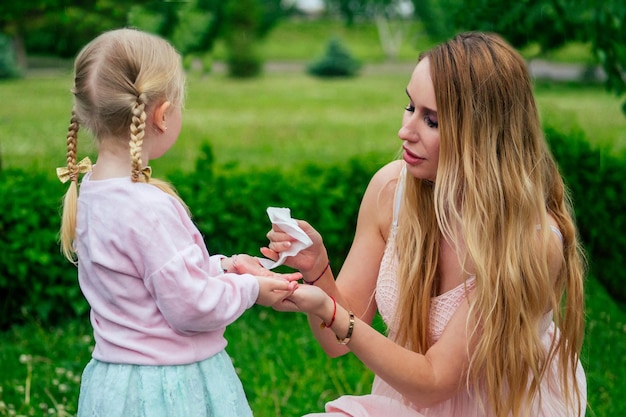 Kleines und süßes blondes Mädchen in einem süßen grauen Kleid zusammen mit einem zahnigen Lächeln schöne Mutter wischt die Hände mit Feuchttüchern im Park vor dem Hintergrund von Bäumen und Grün ab