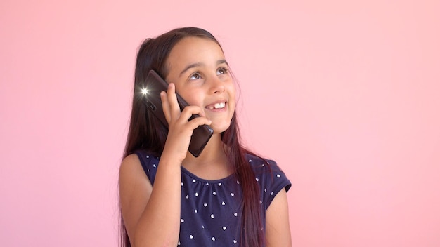 Kleines Vorschulmädchen in einem Kleid, das ein Mobiltelefon hält, isoliert über Rosa