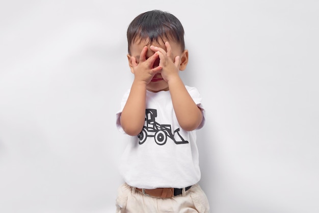 Kleinkind asiatischer Junge 2 Jahre alt trägt ein weißes T-Shirt, das sein Gesicht mit der Hand bedeckt, die auf weißem Hintergrund isoliert ist