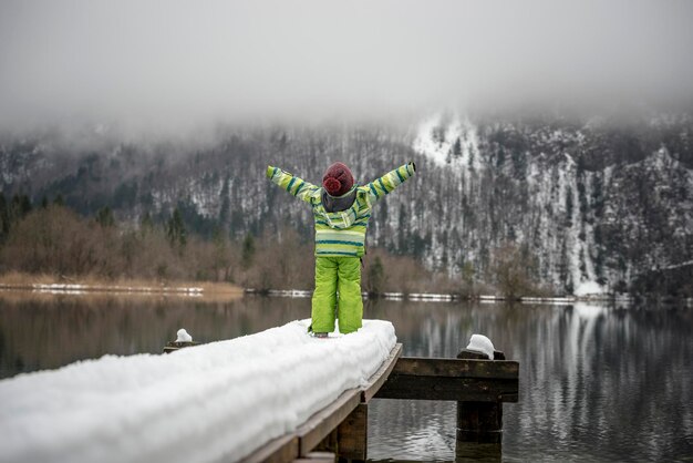 Foto kleinkind im grünen winteranzug steht am ende eines schneebedeckten piers an einem see mit ausgestreckten armen in richtung der wunderschönen winternatur