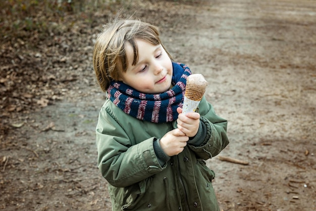 Kleinkind Junge trägt warme Jacke und Schal und isst Schokoladeneis in Waffelkegel mit Vergnügen im Freien bei kaltem Wetter