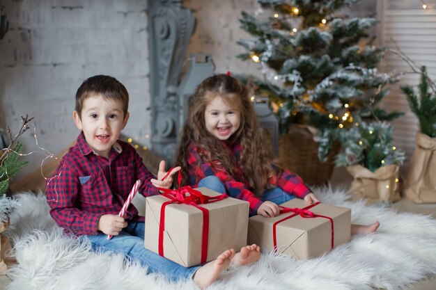 Kleinkind Junge und Mädchen in roten Hemden halten Geschenkboxen in der Nähe von Weihnachtsbaum in hellem Weihnachtsinterieur Süße Weihnachtskinder