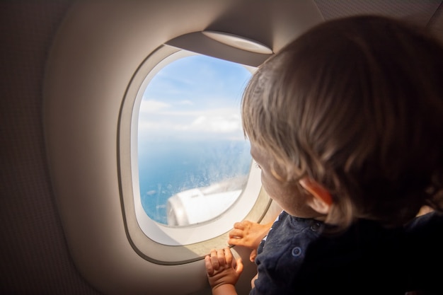 Kleinkind schaut durch das Bullauge eines fliegenden Flugzeugs auf den Boden. mit Kindern reisen