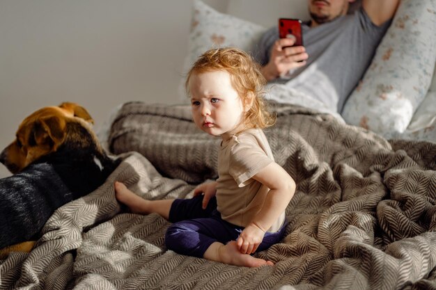 Kleinkind sitzt auf dem Bett mit Vater mit Handy und Haushund im Hintergrund