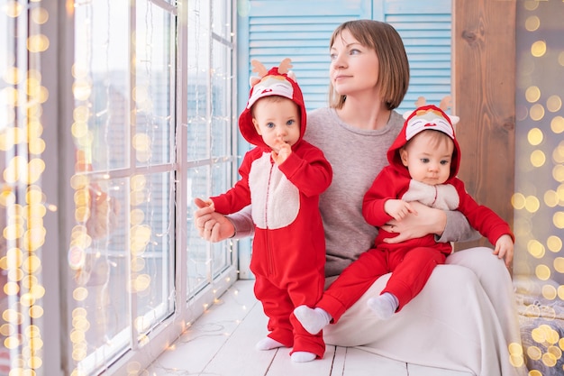 Kleinkind-Zwillinge in roten Rentier-Weihnachtsmann-Kostümen sitzen zu Hause mit ihrer Mutter vor dem Hintergrund eines Fensters mit Girlanden