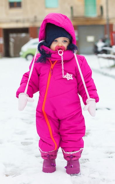 Kleinkindbaby in einer magentaroten Schneeklage, die auf dem Schnee spielt.