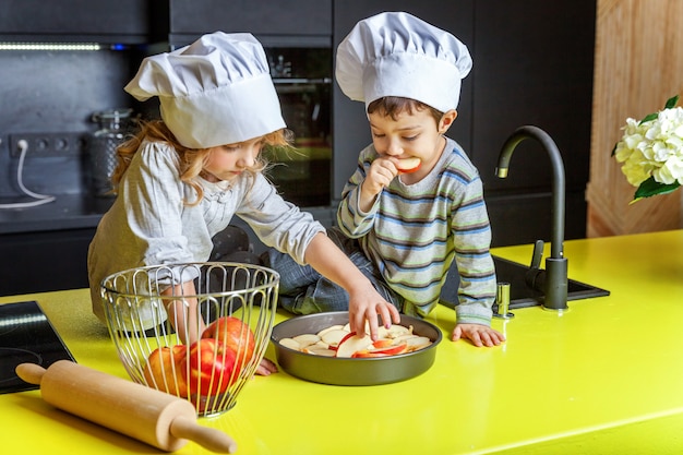 Kleinkinder mädchen und junge mit dem chefhutvorbereiten backen selbst gemachten apfelkuchen in der küche