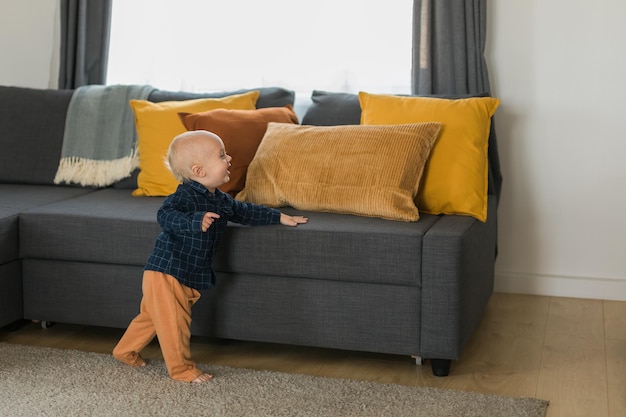 Kleinkindjunge lacht, der Spaß hat, in der Nähe des Sofas im Wohnzimmer zu Hause zu stehen, Kopienraum Entzückendes Baby, das die ersten Schritte allein macht Glückliches Kindheits- und Kinderbetreuungskonzept