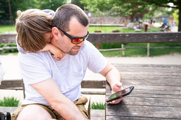 Kleinkindjunge umarmt seinen Vater fest und schaut im Sommer auf das Smartphone im Park Kind stört seinen Vater, der Arbeit und Kinder kombiniert