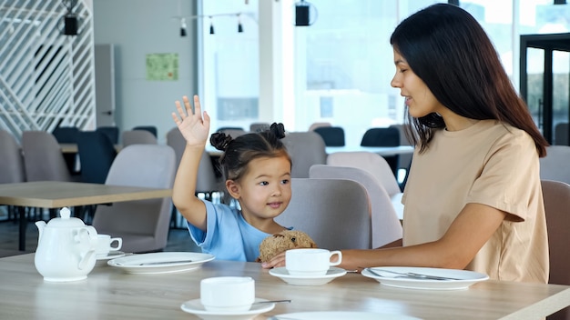 Kleinkindmädchen hebt die Hand mit einem Lächeln, das von Mama im Café sitzt