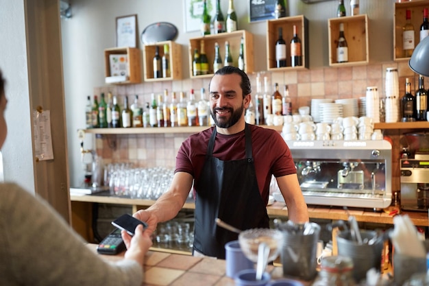 Kleinunternehmen, Menschen und Servicekonzept - glücklicher Mann oder Kellner in Schürze mit Kartenleser und Kunde mit Smartphone, der an der Bar eines Cafés bezahlt