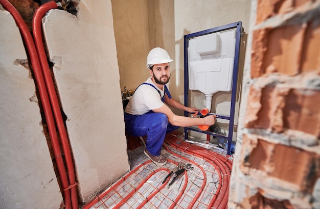 Klempner, der in einer unfertigen Wohnung arbeitet und einen Spülkasten installiert, blickt in die Kamera. Rote Rohre an der Fußbodenheizung. Konzept der Gebäudesanierung, Sanitärarbeiten und Hausrenovierung