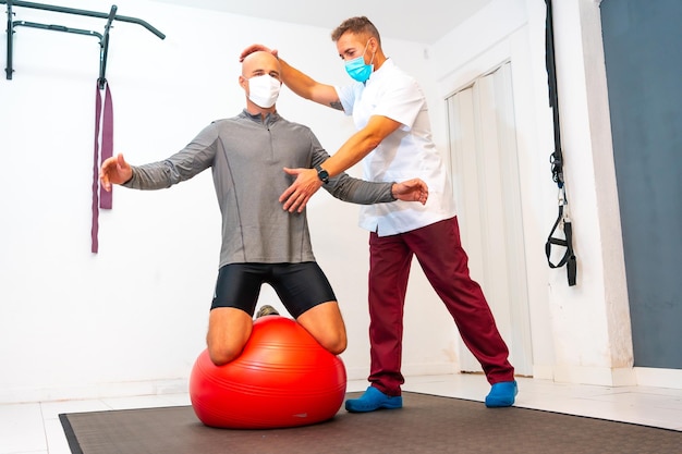 Klient balanciert auf einem roten Ball, unterstützt von einem Physiotherapeuten mit Gesichtsmaske Physiotherapie mit Schutzmaßnahmen für die Coronavirus-Pandemie COVID19 Osteopathie-Sport-Quiromassage