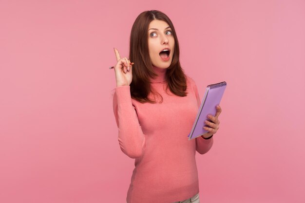 Kluge, erstaunte Frau mit braunen Haaren in rosafarbenem Pullover, die mit dem Finger nach oben zeigt und die Idee hat, in den Notizblock zu schreiben und an einem Buch zu arbeiten. Studioaufnahme im Innenbereich isoliert auf rosa Hintergrund
