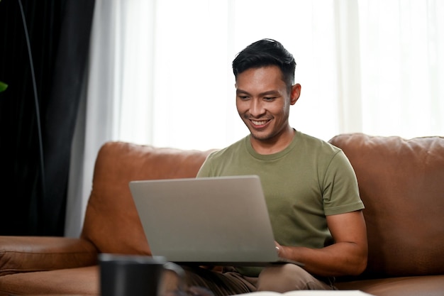 Kluger asiatischer Mann, der einen Laptop auf einem Sofa in seinem Wohnzimmer benutzt, arbeitet von zu Hause aus in der Freizeit