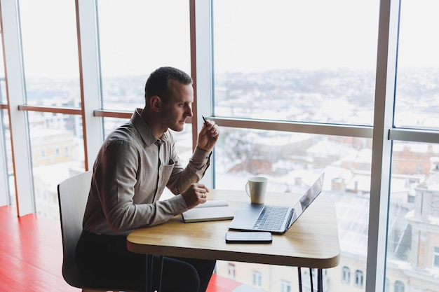 Kluger junger Mann, der auf einer Website nach Informationen sucht und als Freiberufler an der Entwicklung eines Grafikprojekts auf einem modernen Laptop arbeitet, der eine drahtlose Internetverbindung in einem Café-Interieur verwendet