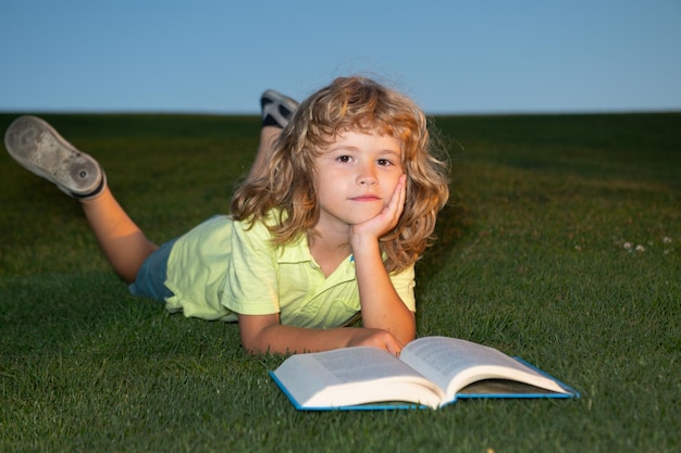 Kluger Kinderjunge, der ein Buch liest, das auf Gras auf Gras- und Himmelhintergrund mit Kopienraum legt