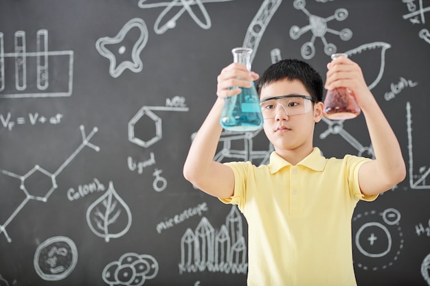 Kluger vietnamesischer Schüler in der Schutzbrille, die Becher mit bunten Flüssigkeiten in seinen Händen betrachtet, Tafel mit Zeichnungen