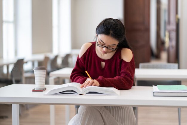 Kluges asiatisches Mädchen, das Umrisse schreibt, während es sich auf die Prüfung im Lesesaal oder Klassenzimmer vorbereitet