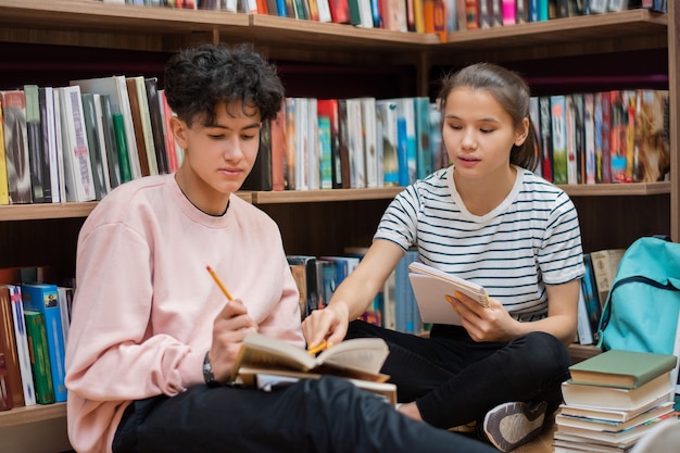 Kluges selbstbewusstes Mädchen, das auf Seite des Buches zeigt, das von ihrer Klassenkameradin während der Vorbereitung für das Seminar in der Universitätsbibliothek gehalten wird