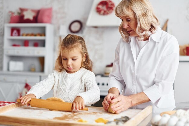 Knetet den Teig Ältere Großmutter mit ihrer kleinen Enkelin kocht Süßigkeiten für Weihnachten auf der Küche