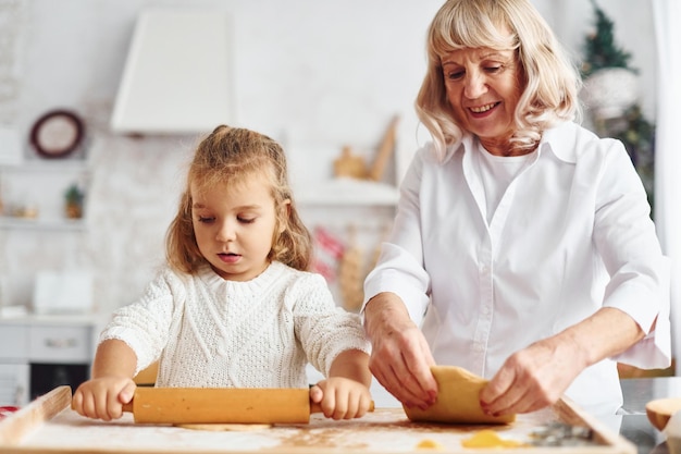 Knetet den Teig Ältere Großmutter mit ihrer kleinen Enkelin kocht Süßigkeiten für Weihnachten auf der Küche