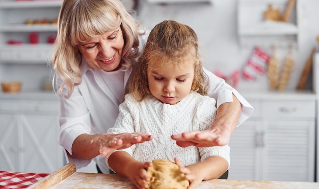 Knetet den Teig Ältere Großmutter mit ihrer kleinen Enkelin kocht Süßigkeiten für Weihnachten auf der Küche