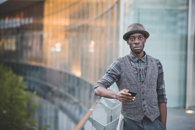 Kniefigur des jungen hübschen Afromannes, der einen Smartphone lehnt auf einem Handlauf hält