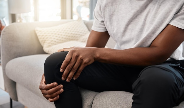 Knieschmerzen Hand eines Mannes und Verletzung beim Sitzen auf dem heimischen Sofa im Wohnzimmer, um sich auszuruhen und zu entspannen Schmerzhaftes Beingelenk und afroamerikanischer Mann, der seine Knie für eine Muskelzerrung berührt