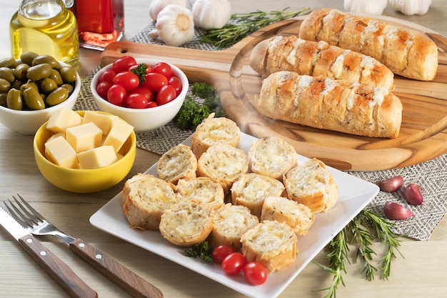 Knoblauchbrot in weißer quadratischer Platte auf dem Tisch mit Käse, Rosmarin, Oliven und Kirschtomaten.