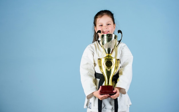 Knockout energía y actividad para niños niña ganadora en ropa deportiva gi niña pequeña con copa de campeón arte marcial practicando Kung Fu éxito deportivo infantil feliz en un solo espacio de copia de combate