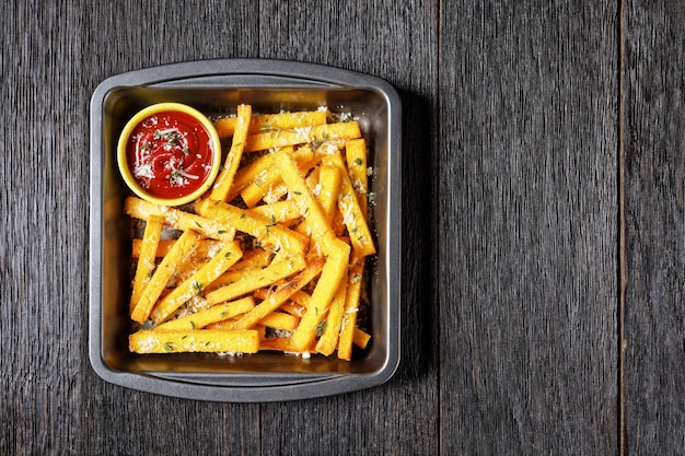 Knusprig gebackene Polenta-Pommes mit Parmesan, Thymian und Gewürzen in einer Auflaufform mit Tomatensauce auf einem rustikalen Holztisch, italienische Küche