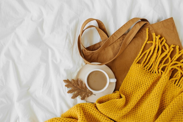 Öko-Tasche aus Baumwolle mit Tasse Kaffee und gelbem Strickschal auf dem Bett. Herbst-Konzept.