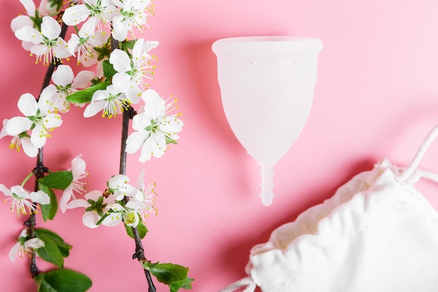 Öko-weiße Menstruationstasse und Baumwollbeutel auf rosa Hintergrund Kirschbaumzweige im Frühling mit weißen Blumen Ökologie- und Recyclingkonzept Zero Waste Women39s Hygiene Menstruation kritische Tage