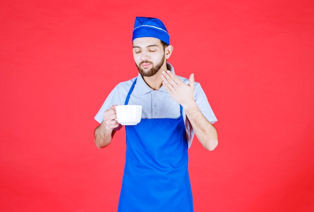 Koch in blauer Schürze, der eine weiße Keramiktasse hält und das Getränk riecht.