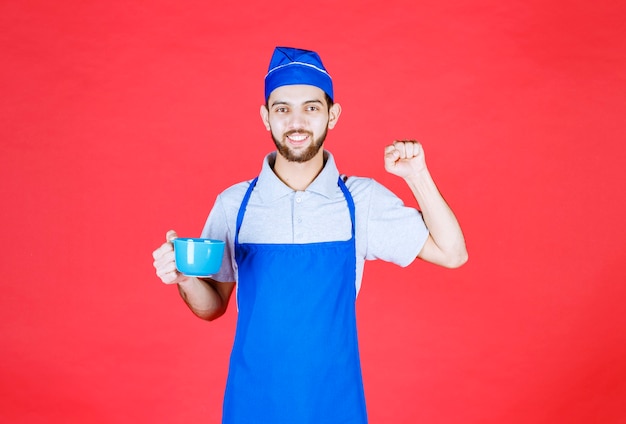 Koch in blauer Schürze, die eine blaue Keramikschale hält und seine Faust zeigt.