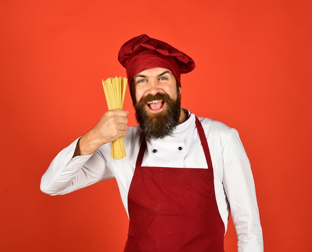 Koch mit fröhlichem Gesicht in burgunderrotem Hut und Schürze hält trockene Nudeln Koch mit Haufen Spaghetti Mann oder Hipster mit Bart hält Makkaroni auf rotem Hintergrund Italienisches Restaurantkonzept