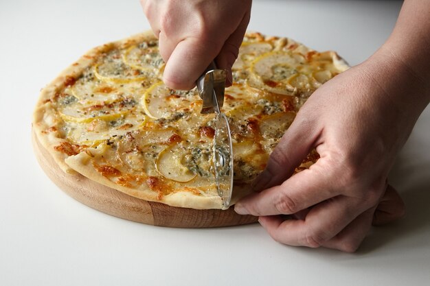 Koch schneidet Birnenfruchtpizza mit Messer auf Holzbrett auf weißer Tischnahaufnahme