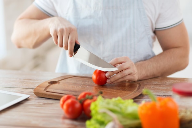 Koch- und Heimkonzept - Nahaufnahme der männlichen Hand, die Tomaten auf Schneidebrett mit scharfem Messer schneidet