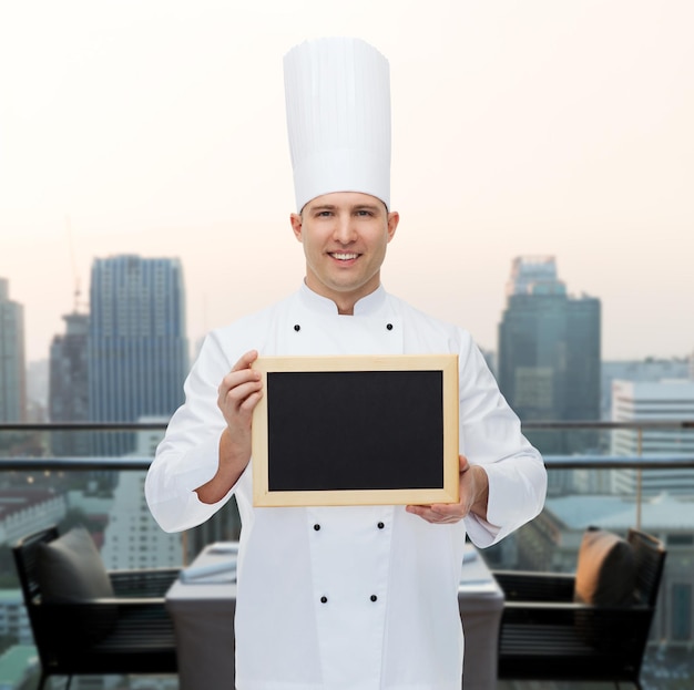 Kochen, Beruf, Werbung und People-Konzept - glücklicher männlicher Koch zeigt und hält leere Menütafel über dem Hintergrund der Stadtrestaurant-Lounge